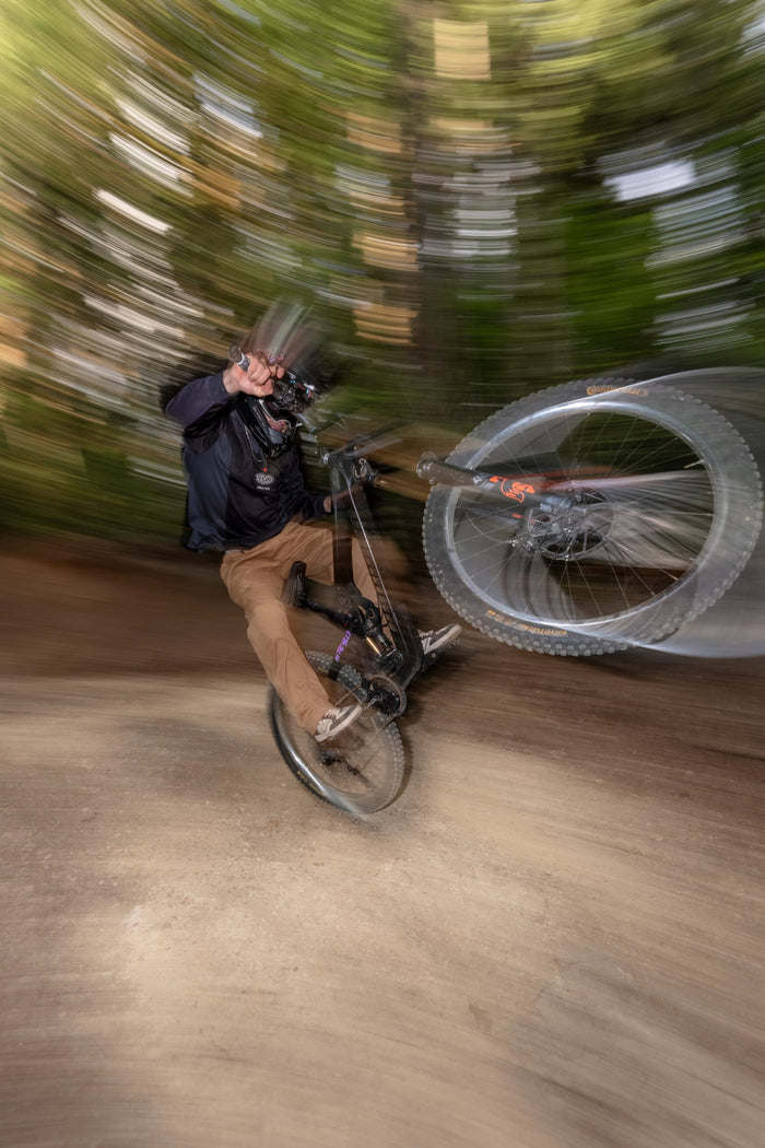 A person wearing the 686 Men's 92 Turbo LS Tech Tee by 686 performs a wheelie on a mountain bike along a forest trail, with motion blur highlighting speed and movement.