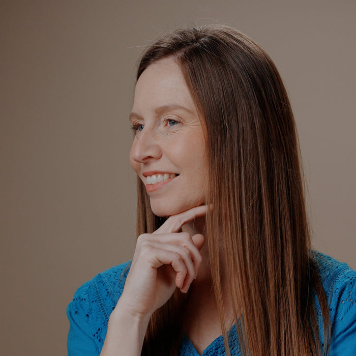 A woman with long brown hair wearing a textured blue top, posing with her hand on her chin against a solid warm brown background.