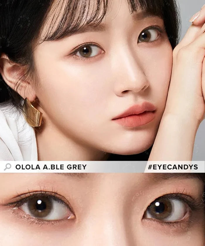 A close-up of a woman with fair skin and straight black hair rests her face on her hand, showcasing Olola Able Grey contact lenses by Olola for dark eyes. The image features the lens detail with text: “Olola Able Grey” and “#EYECANDYS.”.