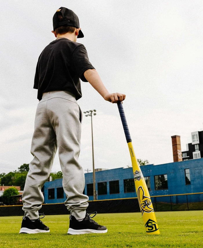 A boy wearing a black cap, black shirt, and gray pants stands on grass holding a Louisville Slugger 2026 Savannah Bananas (-12.5) 2 1/4″ USA T-Ball Baseball Bat (WBL4005010), facing away. A blue building and trees are visible in the background.