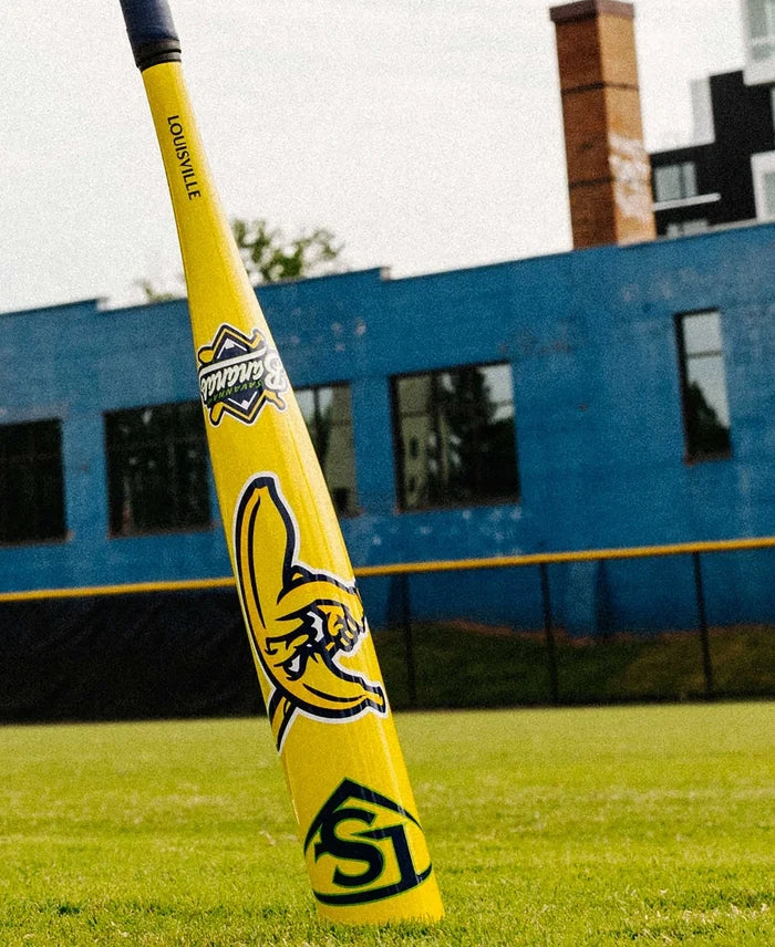 A 2026 Louisville Slugger Savannah Bananas (-12.5) 2 1/4″ USA T-Ball Baseball Bat (WBL4005010) from Louisville Slugger, featuring bright yellow coloring and logos, stands upright on green grass in front of a blue building and a black-and-yellow fence on a cloudy day, evoking the playful style of the Savannah Bananas tee ball bat.