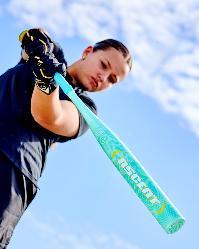 Dressed in black sportswear and gloves, a player grips the 2027 Louisville Slugger Ascent (-12) Fastpitch Softball Bat (WBL4280010), poised to swing beneath a blue sky dotted with white clouds.