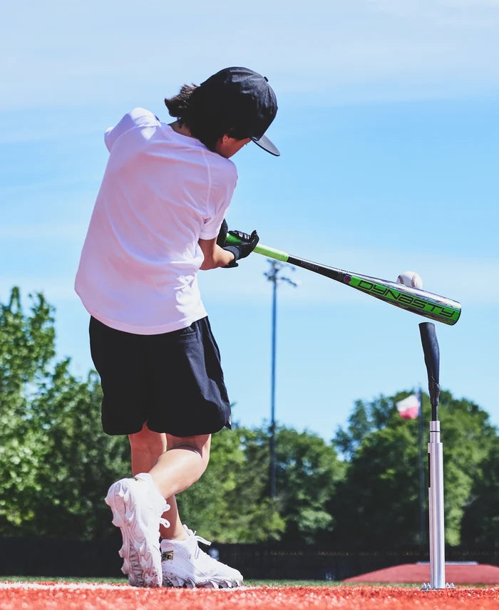 A young person in a white shirt and shorts swings the 2027 Louisville Slugger Dynasty (-11) 2 5/8″ USA Baseball Bat (WL2700101) with a balanced swing weight at a baseball on a tee, outdoors on a sunny day with trees and a blue sky in the background.