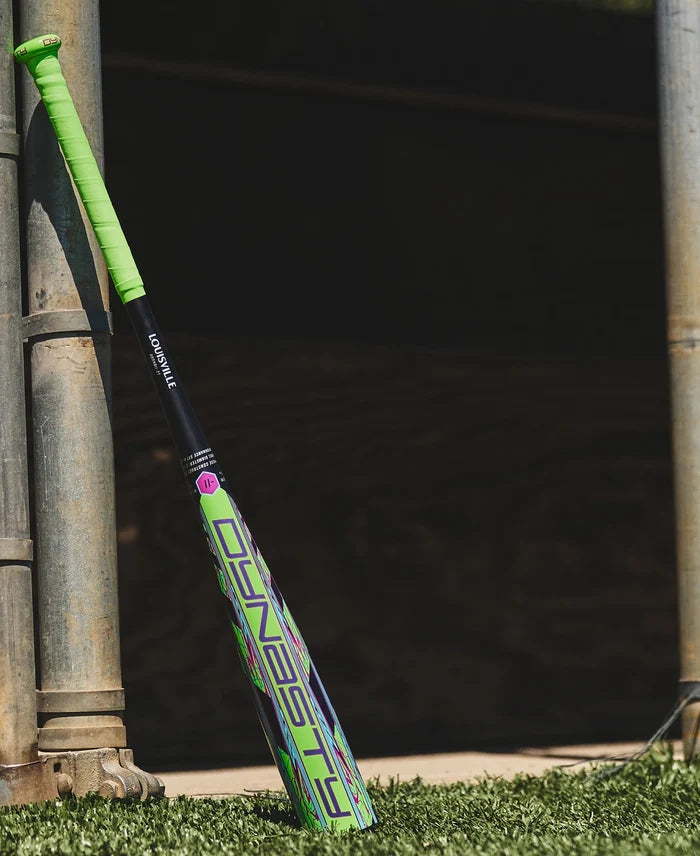 A neon green and black Louisville Slugger 2027 Dynasty (-11) 2 5/8″ USA Baseball Bat (model WL2700101) leans against a metal pole on green artificial turf, next to a wooden wall in sunlight. Engineered as a one-piece alloy bat with balanced swing weight, it’s perfect for your next game.