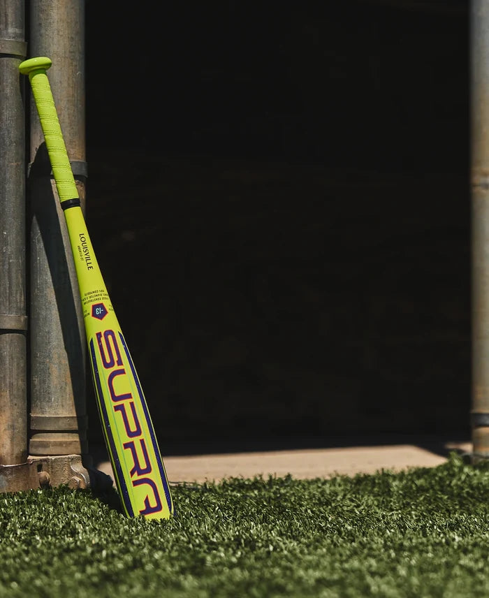 A neon yellow Louisville Slugger 2027 Supra (-13) 2 5/8″ USA T-Ball Baseball Bat (WBL4266010), featuring a green grip, leans against metal poles on artificial turf, set against a dark, shadowy background.