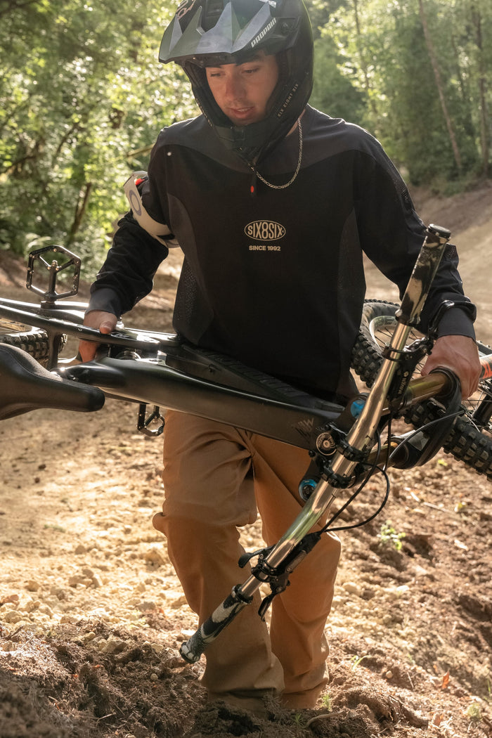 Wearing the 686 Men's 92 Turbo LS Tech Tee, a person in a helmet pushes a mountain bike uphill along a sunlit dirt trail bordered by trees.