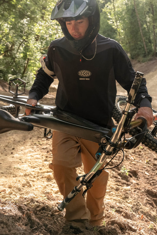 Wearing the 686 Men's 92 Turbo LS Tech Tee, a person in a helmet pushes a mountain bike uphill along a sunlit dirt trail bordered by trees.