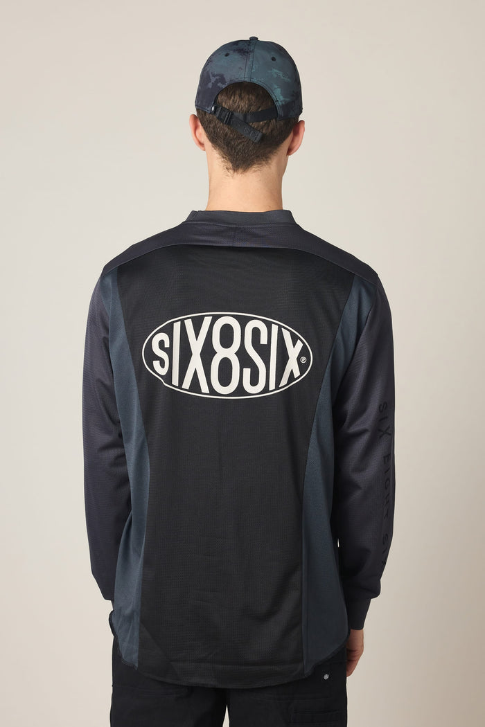 A person wearing a dark cap and the 686 Men's 92 Turbo LS Tech Tee—a black long-sleeve polyester shirt featuring the brand name "686" in a white oval on the back—stands facing away from the camera against a light background.