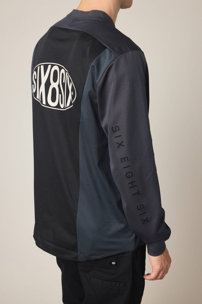 A person wearing the 686 Men's 92 Turbo LS Tech Tee, a dark long-sleeve polyester t-shirt by 686 featuring "SIX8SIX" on the back and "SIX EIGHT SIX" on the sleeve, stands with their back to the camera.