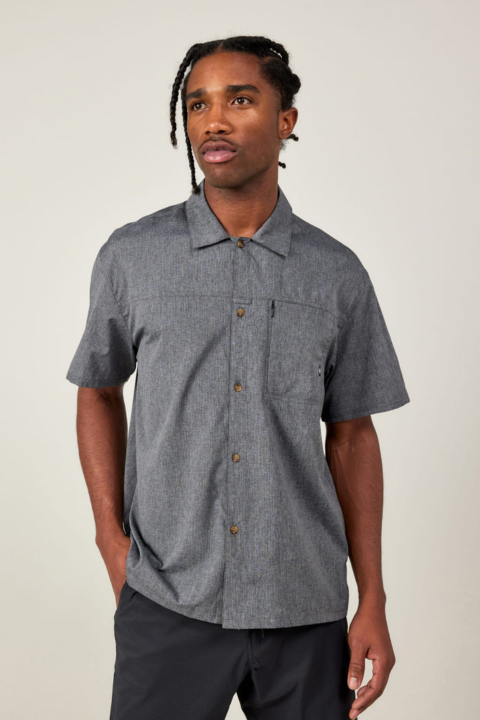 A man with braided hair wears the 686 Mens Canopy Perforated Button Up shirt, featuring short sleeves and a chest pocket, along with black pants. He stands against a plain light background, looking slightly to the side with a neutral expression.