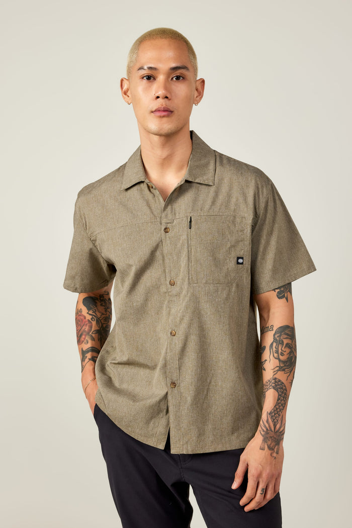 A person with short blond hair and visible tattoos on both arms wears the 686 Mens Canopy Perforated Button Up and dark pants, standing against a plain light background.