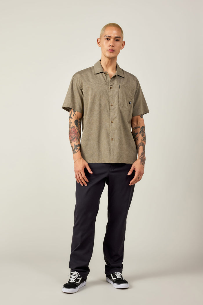 A person with short blond hair and arm tattoos stands facing forward in a 686 Mens Canopy Perforated Button Up, dark pants, and black sneakers against a plain light background.