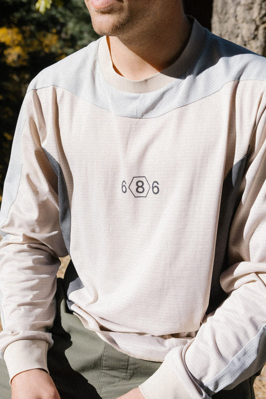 A person is sitting outdoors in sunlight, wearing the 686 Men's Hex Turbo LS Tech Tee—a beige long-sleeve shirt featuring the number 686 and a hexagon logo on the chest. This stylish tee from 686 is made of 100% polyester, providing comfort and durability all day long.