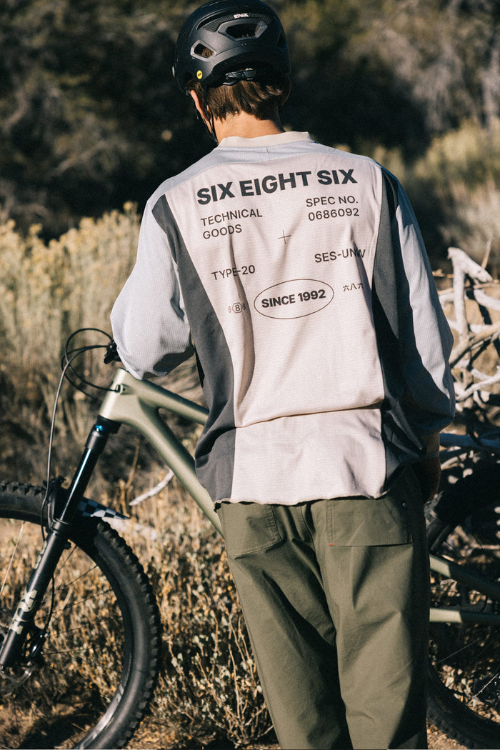 A person wearing a helmet and a 686 Men's Hex Turbo LS Tech Tee made from 100% polyester stands beside a mountain bike outdoors, facing away from the camera amidst dry vegetation.