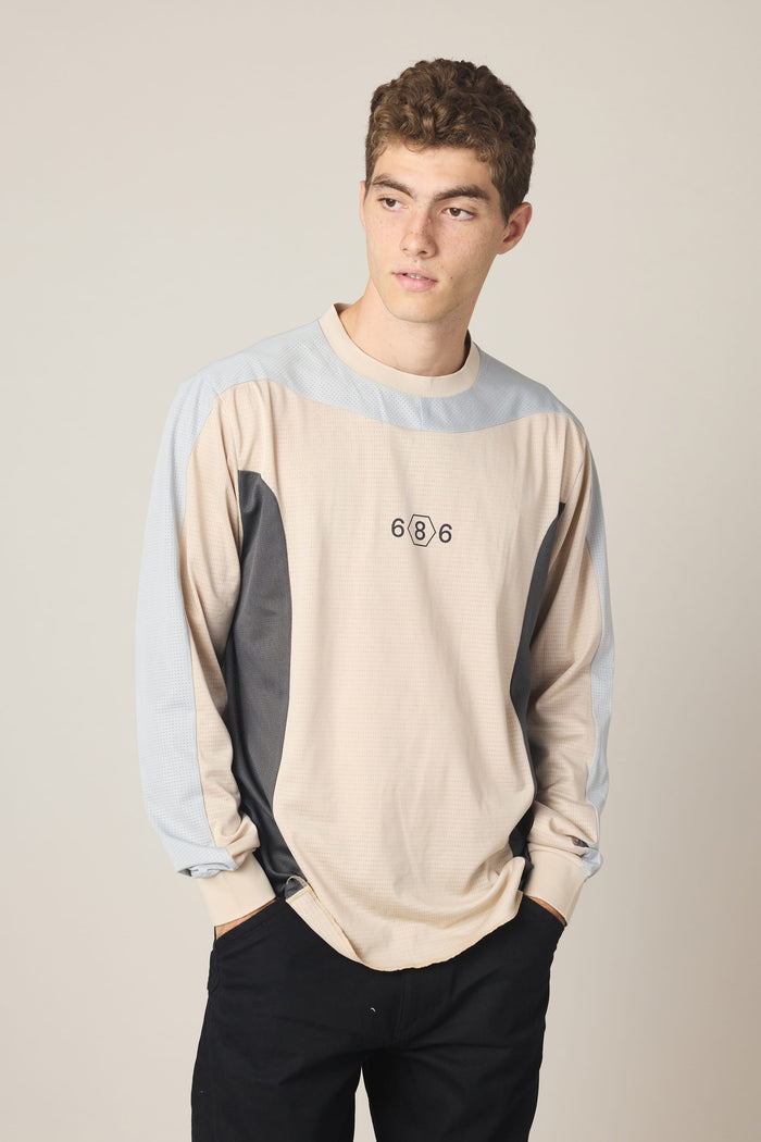 A young man with short curly hair stands against a plain light background with his hands in his pockets, wearing the 686 Men’s Hex Turbo LS Tech Tee in beige and light blue—featuring 686 branding on the chest—and black pants.