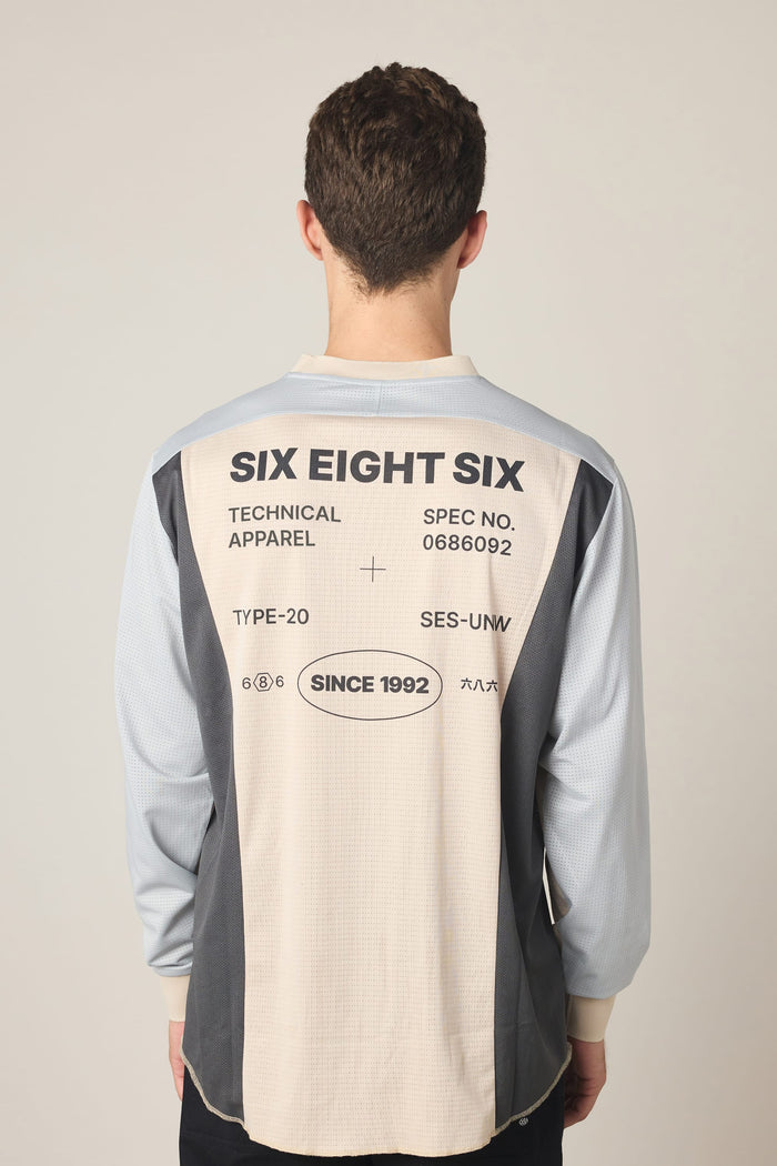 A person faces away, wearing the 686 Men's Hex Turbo LS Tech Tee by 686—a long-sleeve t-shirt featuring SIX EIGHT SIX and technical details printed in black on a beige and gray color-blocked back. Made from 100% polyester, this tee delivers both style and performance.