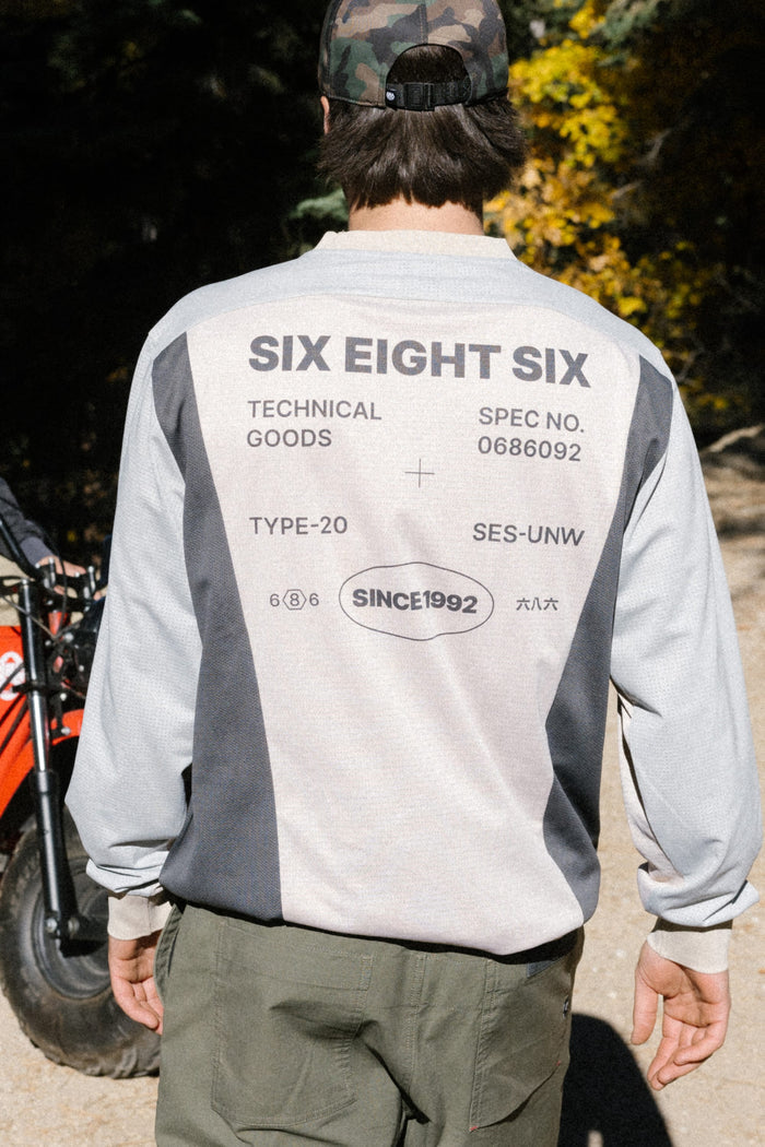 A person wearing a 686 Men's Hex Turbo LS Tech Tee stands outdoors. The long sleeve tee, made from 100% polyester and adorned with various text and graphics, is worn with the person facing away from the camera. Trees and part of a red vehicle can be seen in the background.
