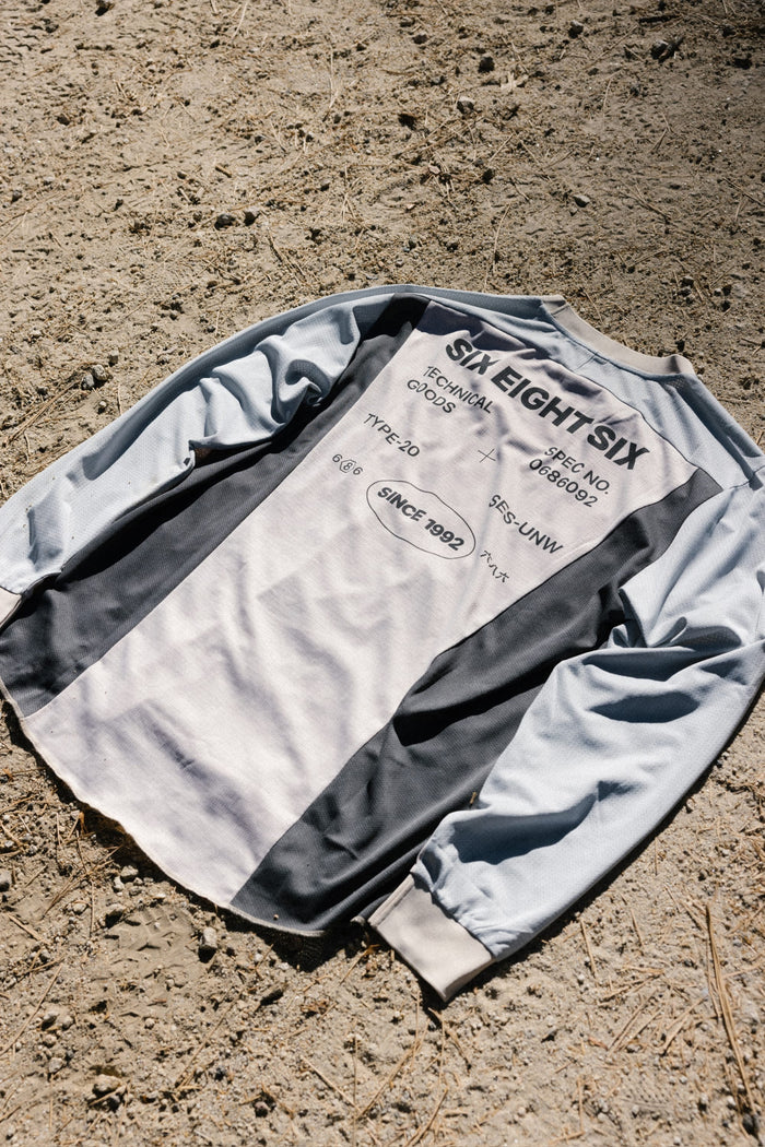 The 686 Men's Hex Turbo LS Tech Tee, featuring black and white panels, lies on dry, rocky ground. This long-sleeve t-shirt from 686 displays printed text such as SIX EIGHT SIX, technical details, and SINCE 1992. Made from 100% polyester for comfort and durability.