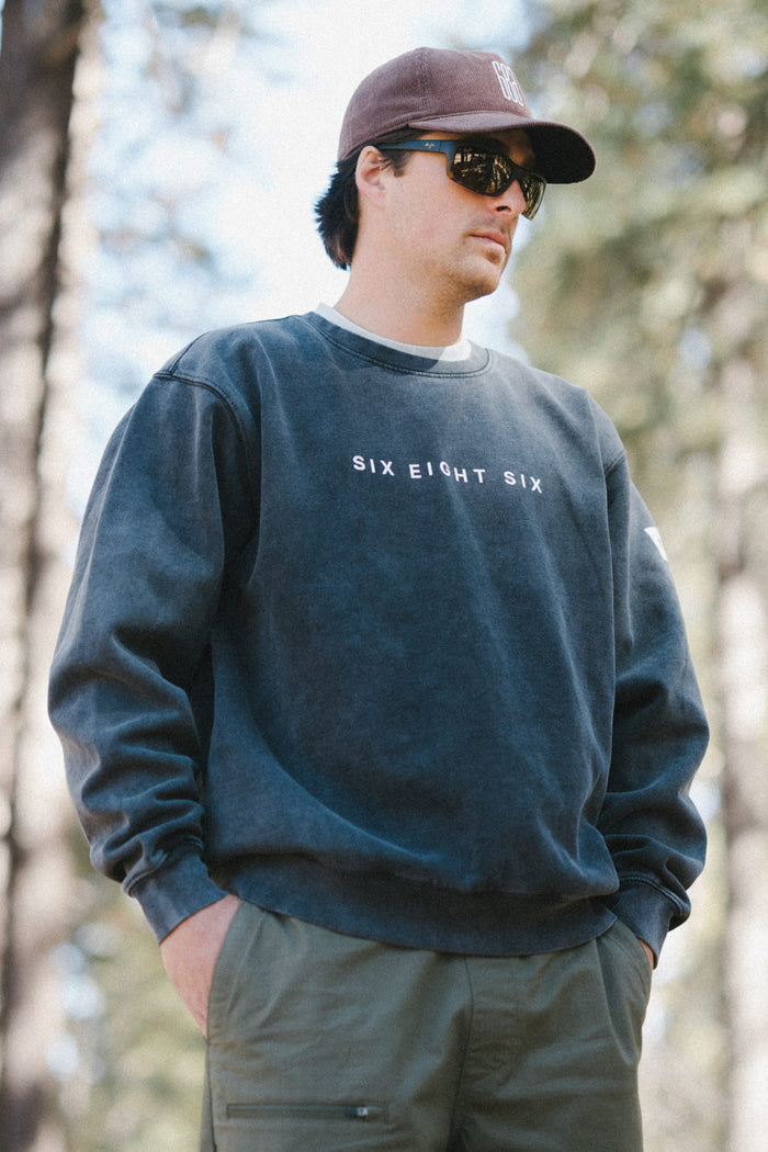 A person stands outdoors in a sunlit forest, wearing sunglasses, a dark cap, and the 686 Men's Mixed Relaxed LS Crewneck sweatshirt layered over a classic men's t-shirt.