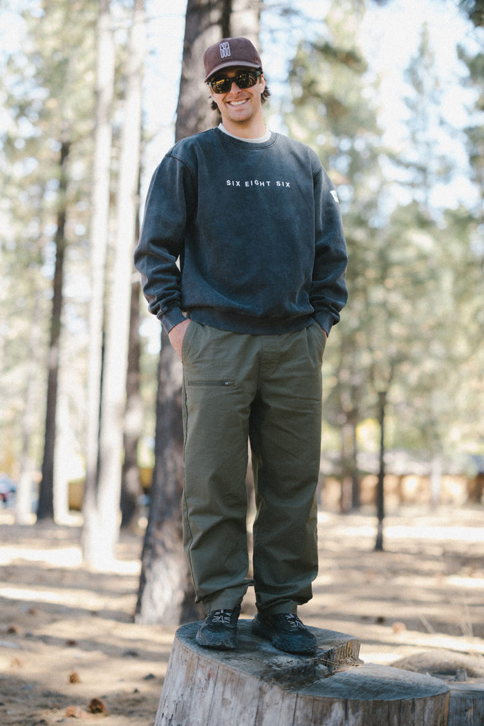 A person wearing sunglasses, a dark 686 Men's Mixed Relaxed LS Crewneck over a classic men's t-shirt, cap, and green pants stands on a tree stump in a forested area, smiling with hands in pockets.