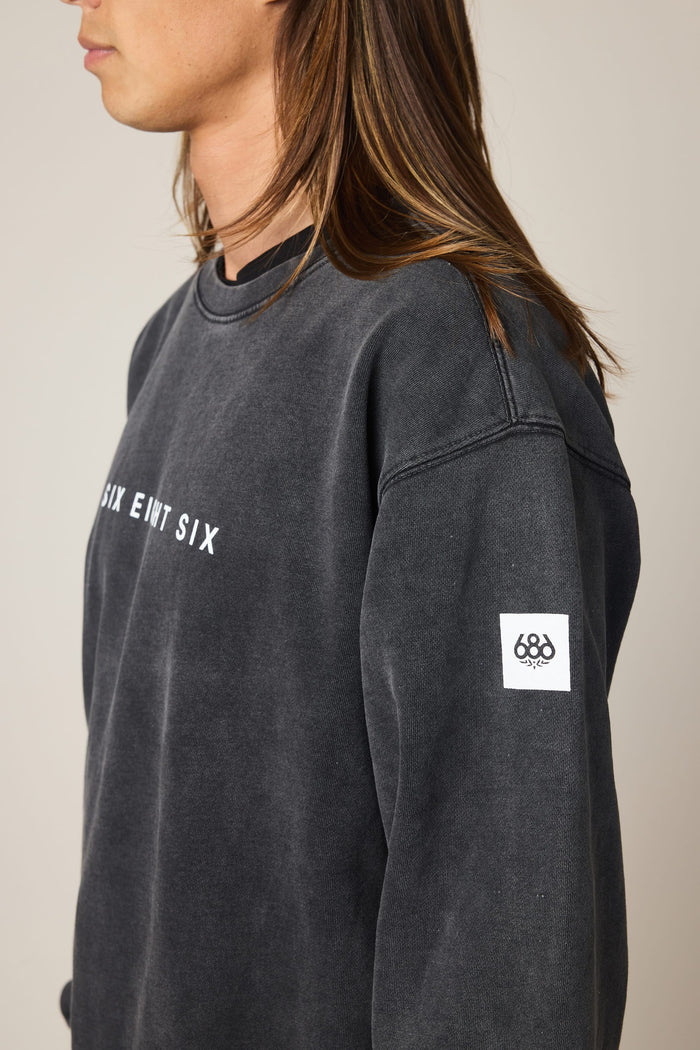 A person with long brown hair is wearing the 686 Men's Mixed Relaxed LS Crewneck, a faded black sweatshirt styled over a classic men's t-shirt. The crewneck features white "SIX EIGHT SIX" text on the chest and a white rectangular "686" patch on the left sleeve. The person is facing left and shown from the chest up.