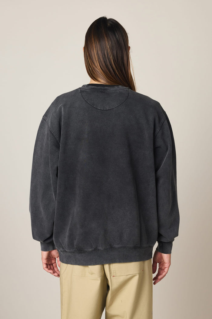 A person with long brown hair, seen from behind against a plain light background, is wearing the 686 Men's Mixed Relaxed LS Crewneck—a loose, faded black 100% cotton sweatshirt from 686—paired with beige pants.