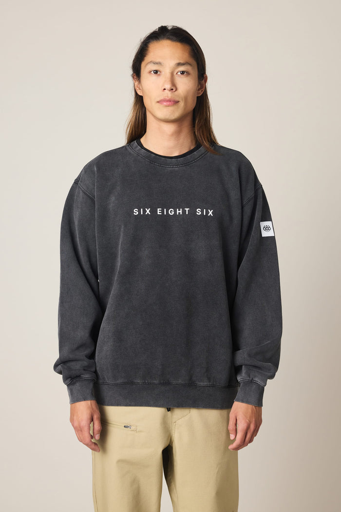 A person with long hair stands facing forward, wearing the 686 Men's Mixed Relaxed LS Crewneck—a faded black sweatshirt from 686. Designed to complement everyday essentials such as a classic cotton t-shirt, this crewneck features “SIX EIGHT SIX” lettering on the front and a small white patch on the left sleeve. The background is plain and neutral.