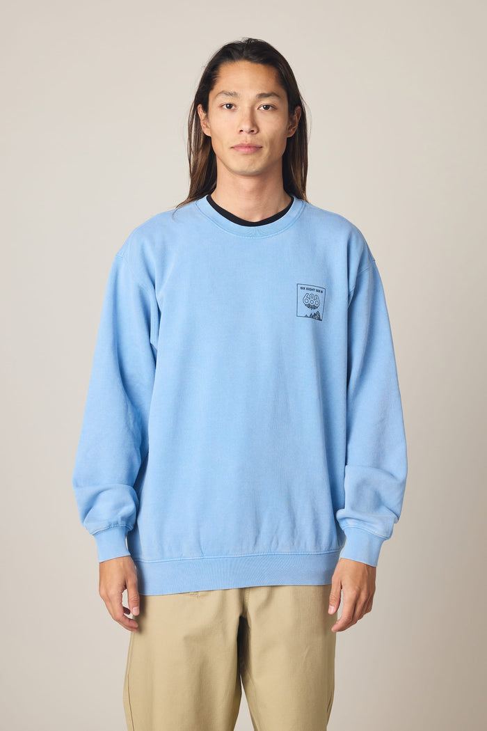 A person with long hair wears the 686 Men's Mixed Relaxed LS Crewneck by 686 in light blue, paired with beige pants, standing against a plain, light background.