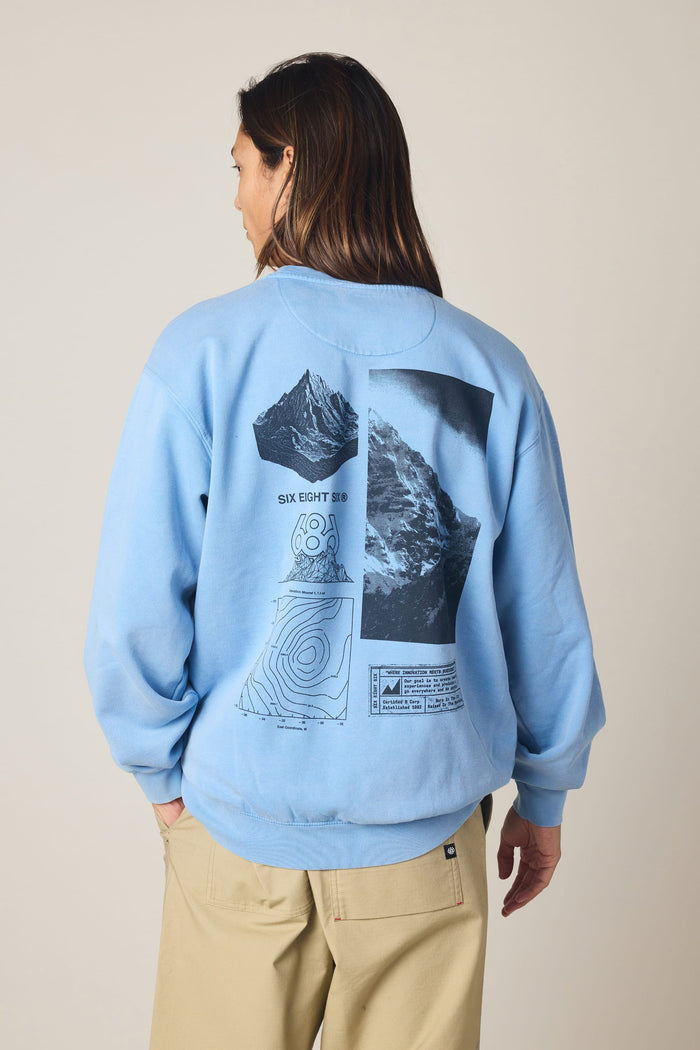 A person wearing the 686 Men's Mixed Relaxed LS Crewneck—a light blue 100% cotton sweatshirt by 686 featuring black-and-white mountain imagery, topographic map graphics, and text on the back—stands against a plain background.