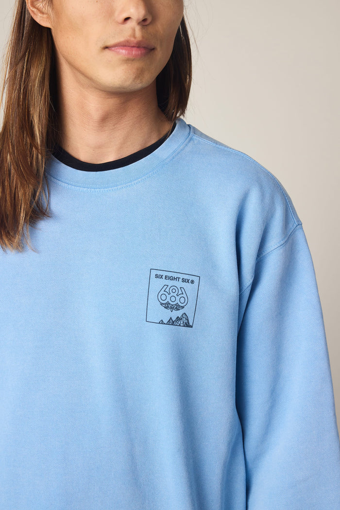 A person with long brown hair is wearing the 686 Men's Mixed Relaxed LS Crewneck—a light blue, 100% cotton sweatshirt by 686 featuring a small embroidered mountain graphic and the text "SIX EIGHT SIX" on the left chest. The background is plain and neutral.
