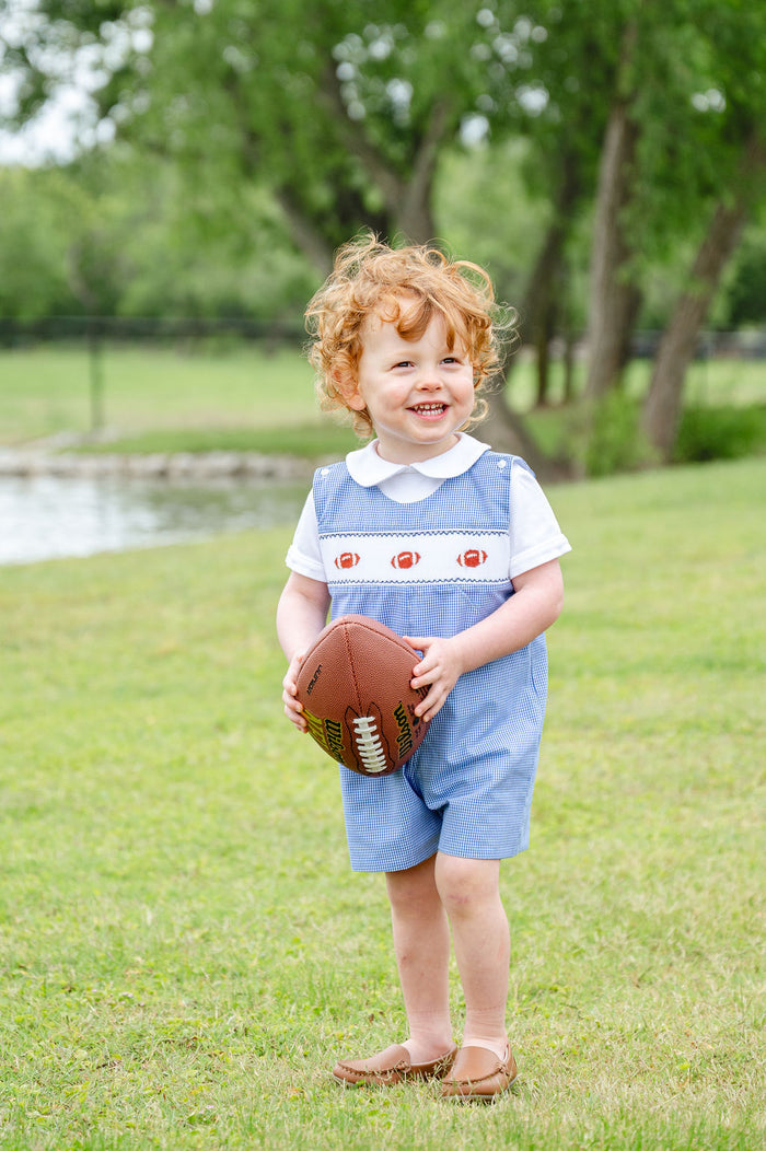 Toddler boy wearing a blue gingham smocked football jonjon with three hand-smocked footballs over a Peter Pan collar shirt, holding a football