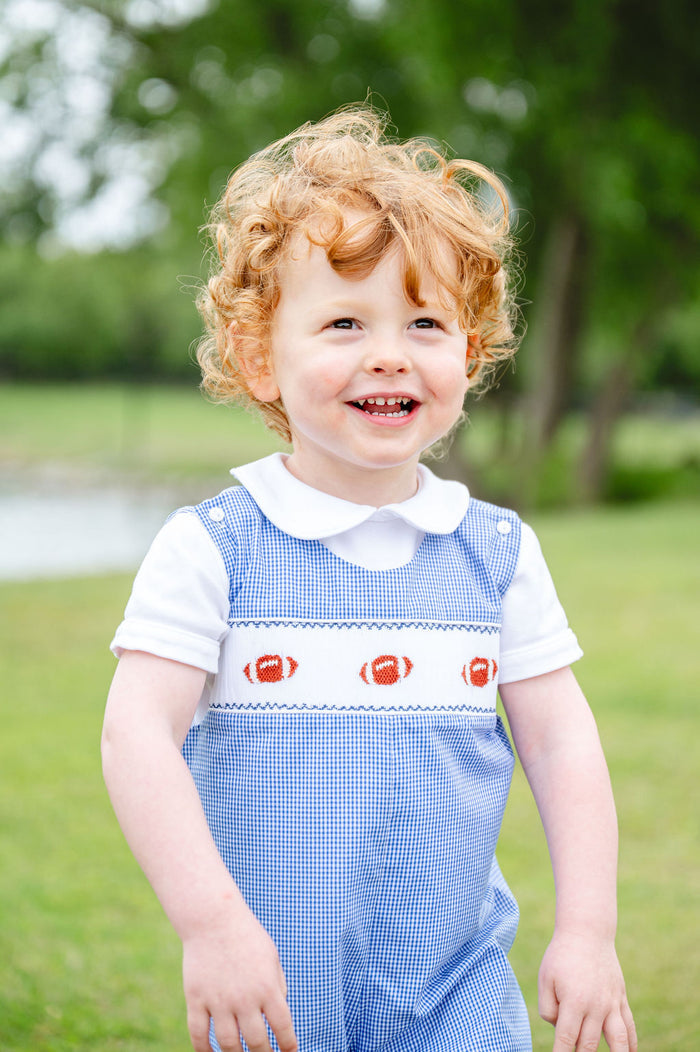 Little boy in a blue gingham jonjon with hand-smocked football panel, standing outdoors holding a football