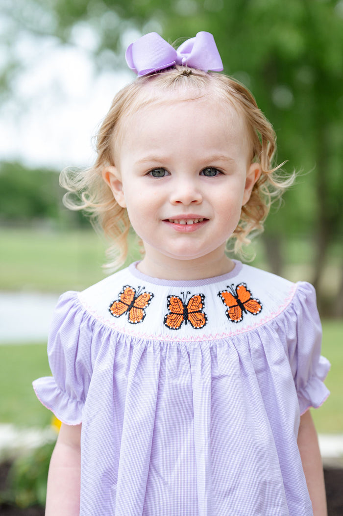 Toddler girl wearing a light purple gingham smocked bloomer set with a hand-smocked neckline of orange monarch butterflies, standing outdoors