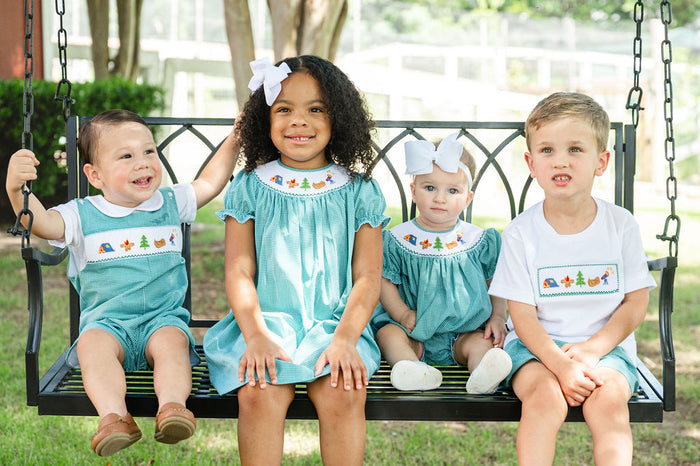 Toddler sitting on porch swing wearing Smocked Camping Jon Jon by Ruth and Ralph, holding a ball