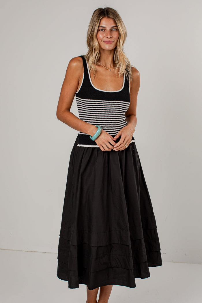 A woman modeling a sleeveless midi dress with a black and white horizontally striped bodice and a full black tiered skirt, paired with tan pointed-toe heels against a plain light gray studio background.