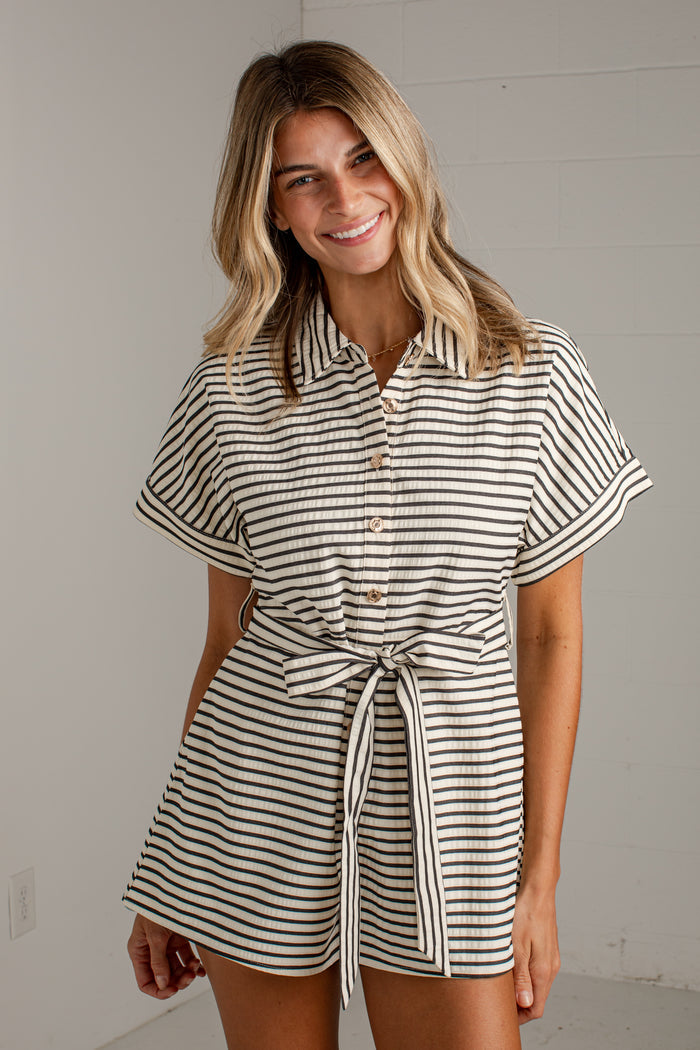 A woman modeling a short-sleeve collared romper with thin black and off-white horizontal stripes, featuring a button front and a self-tie belt at the waist.