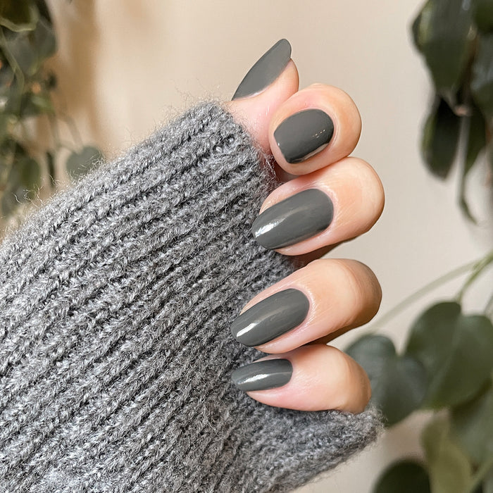 Hand with medium-length nails painted in Is it Smoky in Here? deep charcoal gray gel polish wearing cozy gray sweater sleeve