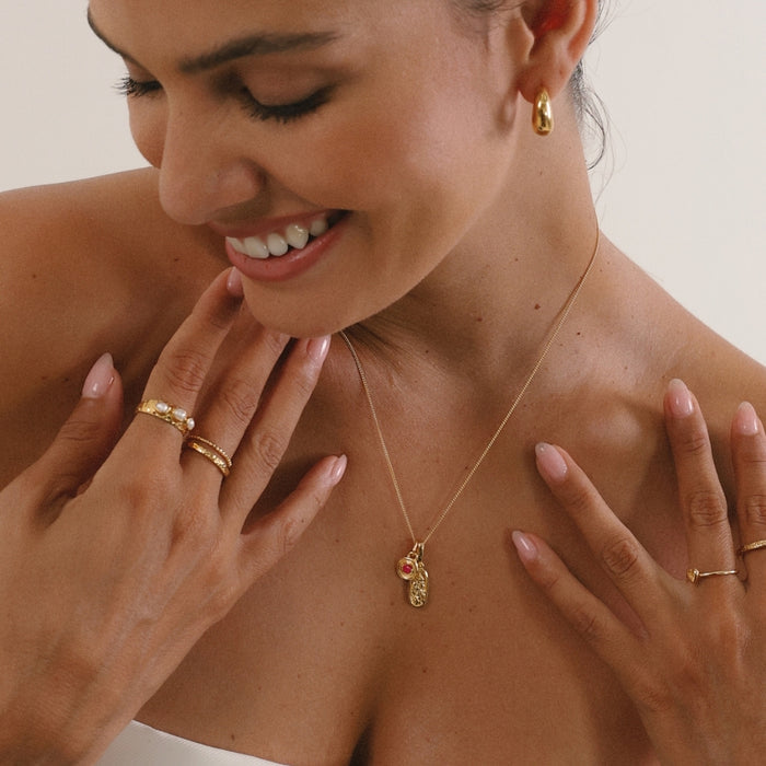 Woman wearing gold jewelry including a necklace, rings, and earrings against a neutral background