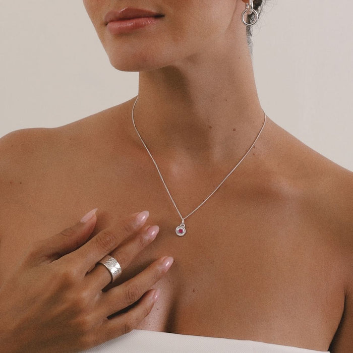 Woman wearing a silver necklace with a pink heart pendant and matching ring on a neutral background