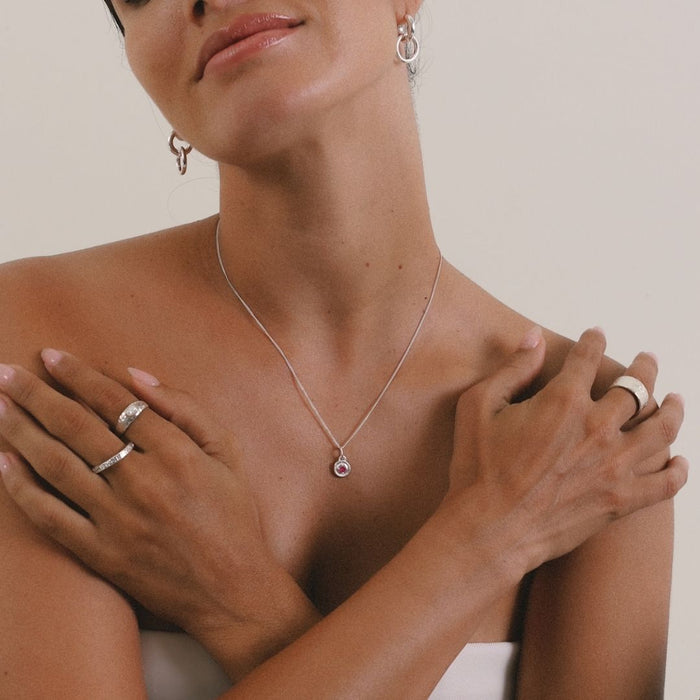 Woman wearing a silver necklace with a pink gemstone, surrounded by jewelry on a neutral background