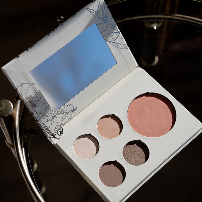 An open makeup palette with a large mirror on the inside lid and five round cosmetic pans in neutral and pink shades, placed on a dark reflective surface.
