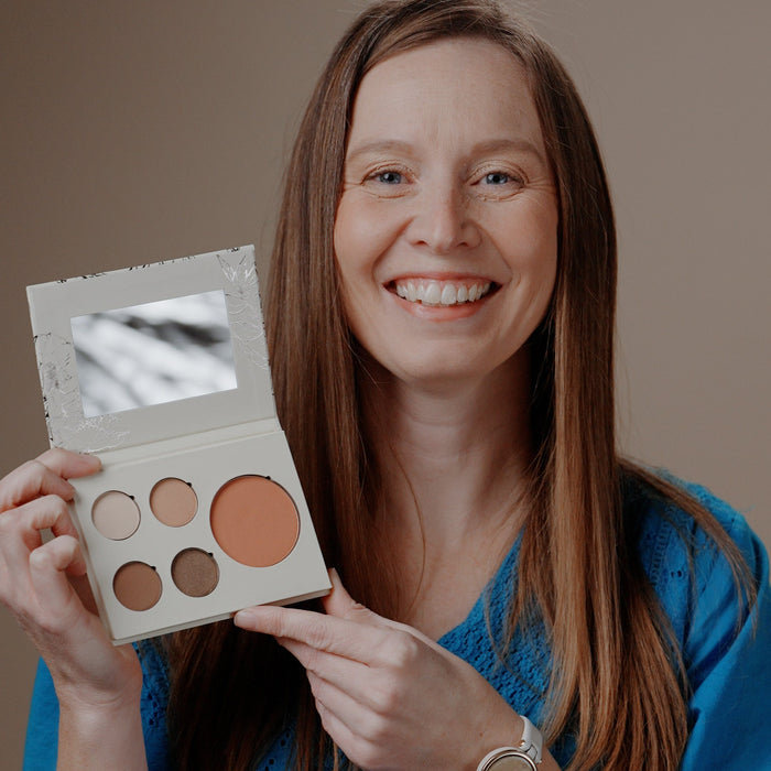 A smiling woman with long brown hair holds up a square makeup palette with a mirror and several warm-toned cream and powder pans against a plain beige background.