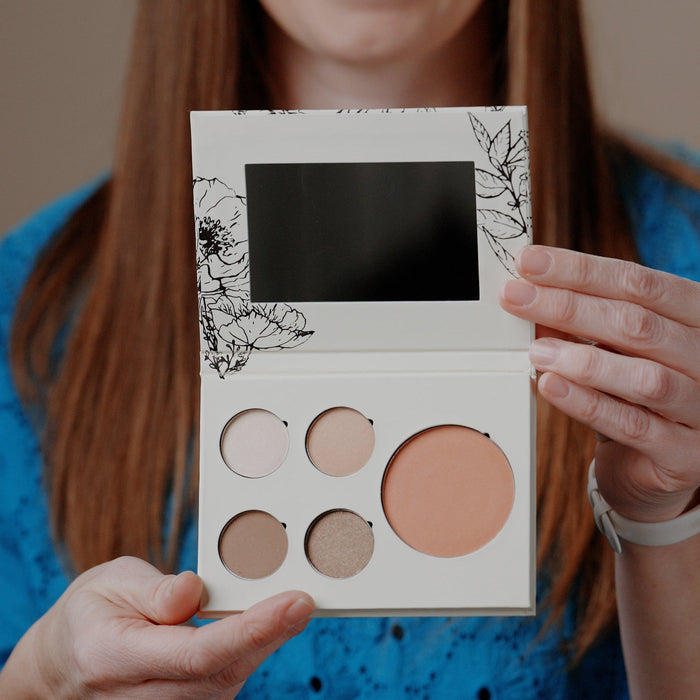 A woman holding an open makeup palette with five circular pans in neutral and peach shades. The palette has a white cover with black floral line art and a black mirror.
