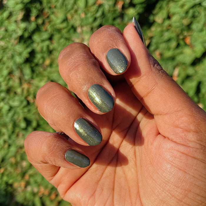 Hand with short nails painted in Martini Chrome showing deep olive green gel polish with golden shimmer finish outdoors