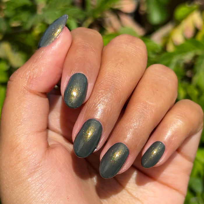Hand with oval nails painted in Martini Chrome showing deep olive green and golden shimmer gel polish manicure outdoors