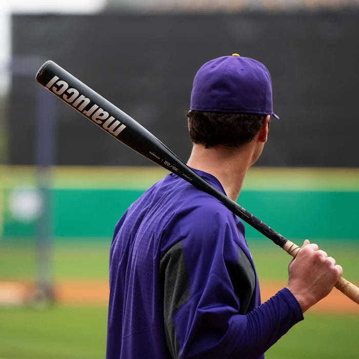 A baseball player in a purple uniform and cap stands on a field, holding a Marucci Aluminum Fungo 35″ Training Bat (MTRBFA) constructed with high-strength alloy over his shoulder, facing away from the camera.