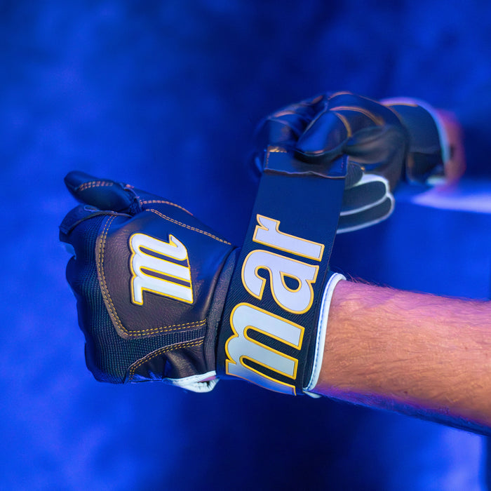 A person wearing durable Marucci Blacksmith V2 Full Wrap Youth Batting Gloves fastens one glove with a large strap displaying “Marucci” in bold yellow letters under blue lighting.