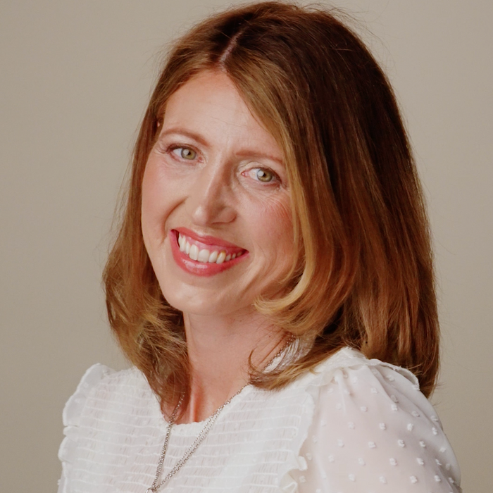 A smiling woman with shoulder-length brown hair wearing a white long-sleeve blouse with subtle polka dots and a delicate gold necklace, photographed against a plain warm beige studio background.