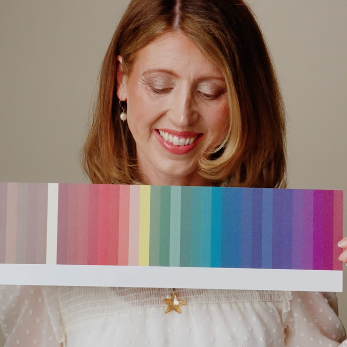 A woman with brown hair smiles while holding a long horizontal color swatch card with vertical stripes in beige, pink, red, orange, yellow, green, blue, and purple. She is wearing a white textured top against a plain beige background.