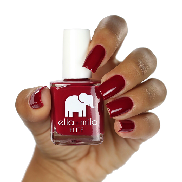Naughty Not Nice deep ruby red nail polish on a medium skin tone, hand holding the bottle